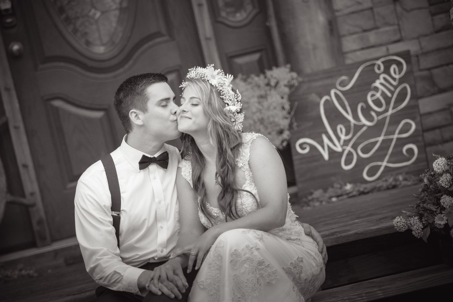 A black and white wedding photo shows a couple sharing a romantic kiss on a bench with decorative neon signage behind them.