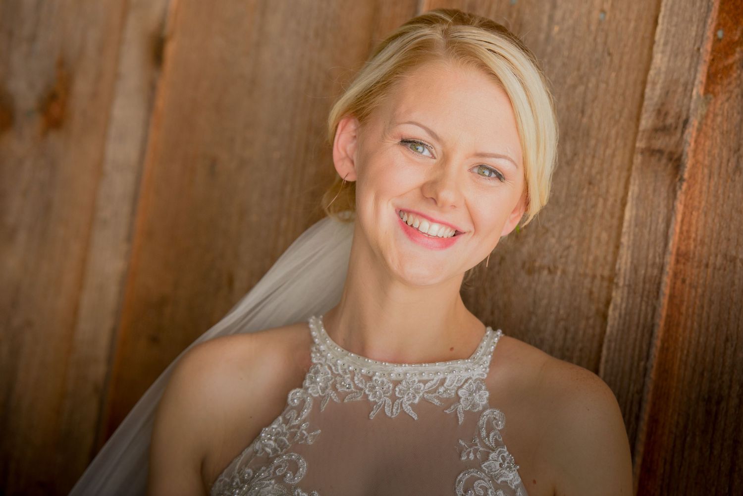 A bride wearing a delicate lace gown smiles warmly while posing against a rustic wooden backdrop by Pigeon Forge Wedding Photographers.