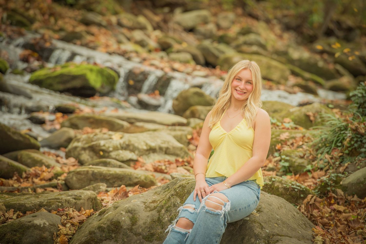 Someone poses on mossy rocks near a cascading waterfall wearing a yellow top and distressed jeans in warm autumn lighting.