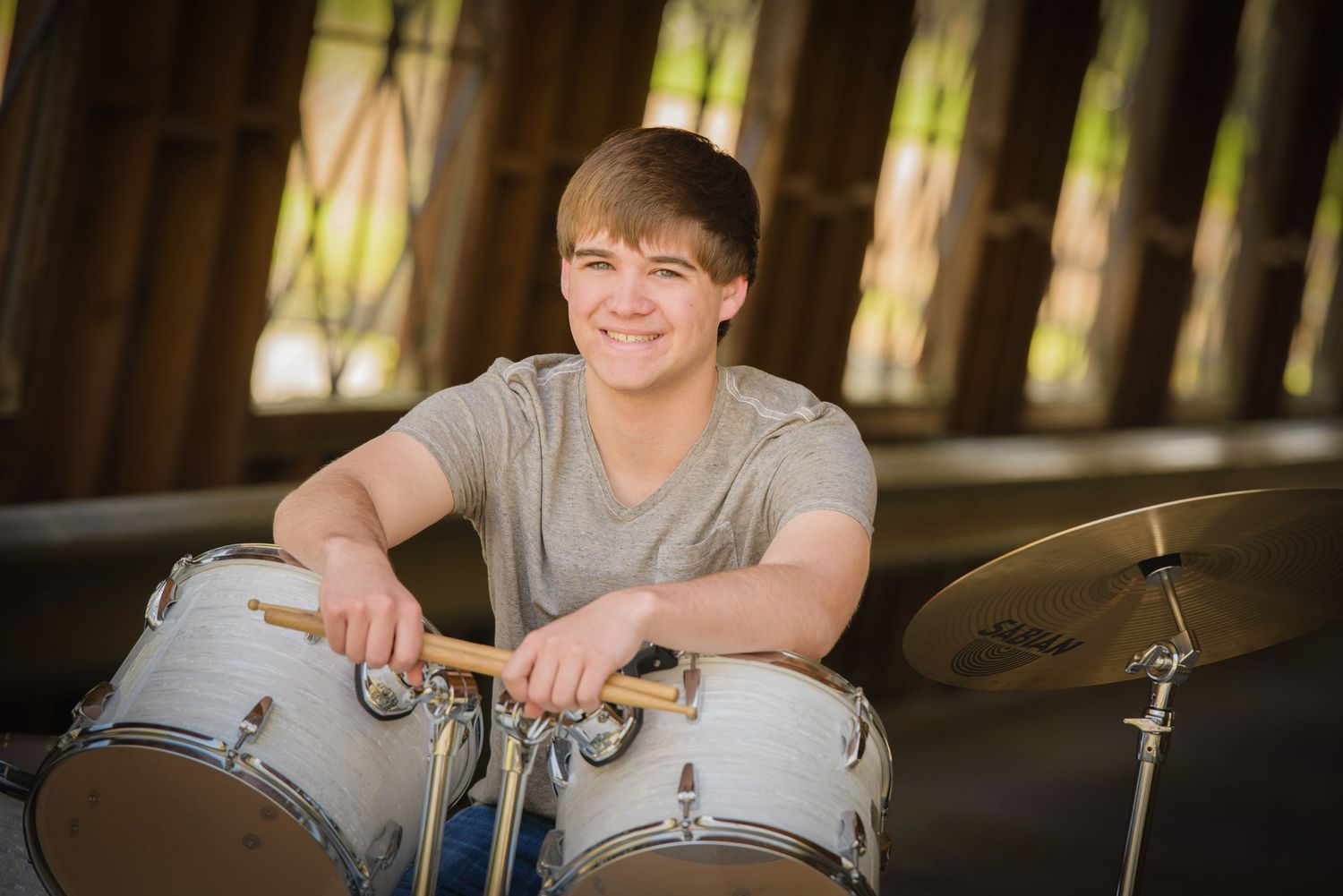Young drummer smiles while seated at white drum kit during outdoor photo session under wooden covered bridge.