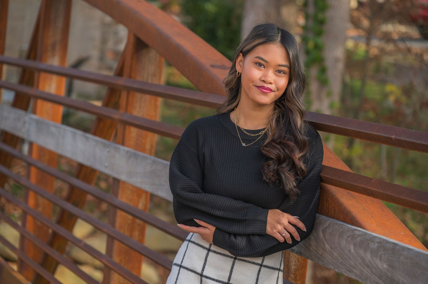 A woman in a black sweater and patterned skirt poses on a wooden deck railing during golden hour.