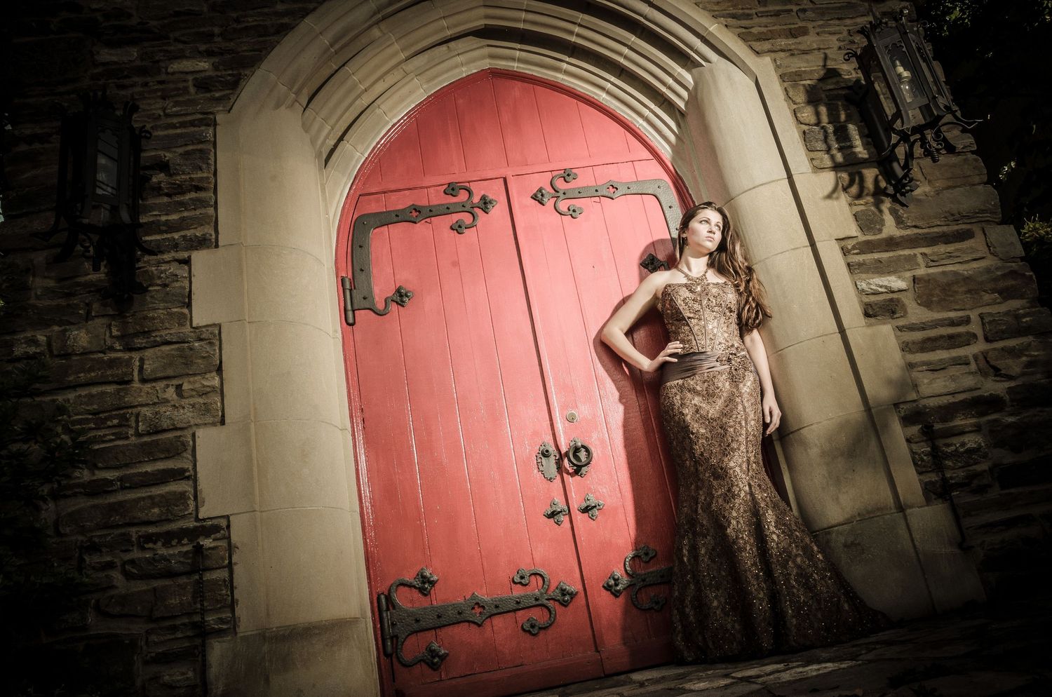 A figure in vintage dress poses against a dramatic pink church door with ornate black metal hinges in gothic archway.