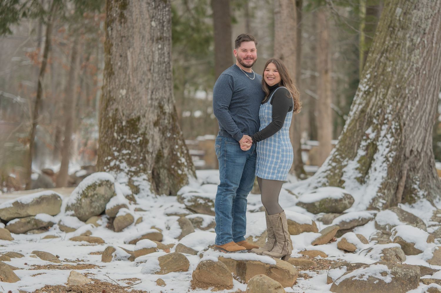 A couple standing together on snowy ground in a winter forest with tall trees and rocks around them by Pigeon Forge Photographer tn.