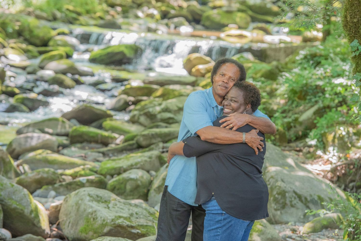 Two people embrace in a tender hug while standing near a scenic rocky waterfall stream in a lush forest setting by Gatlinburg Photographers.