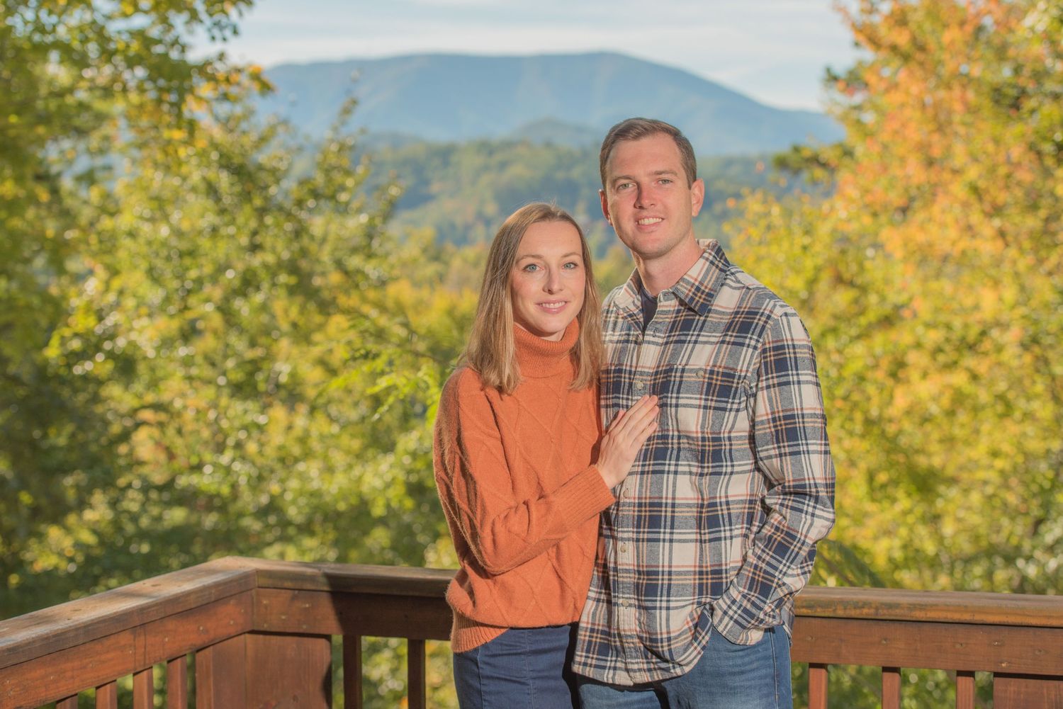 A couple poses on a deck during autumn with mountains and yellow fall foliage in the background by Townsend TN Photographers.
