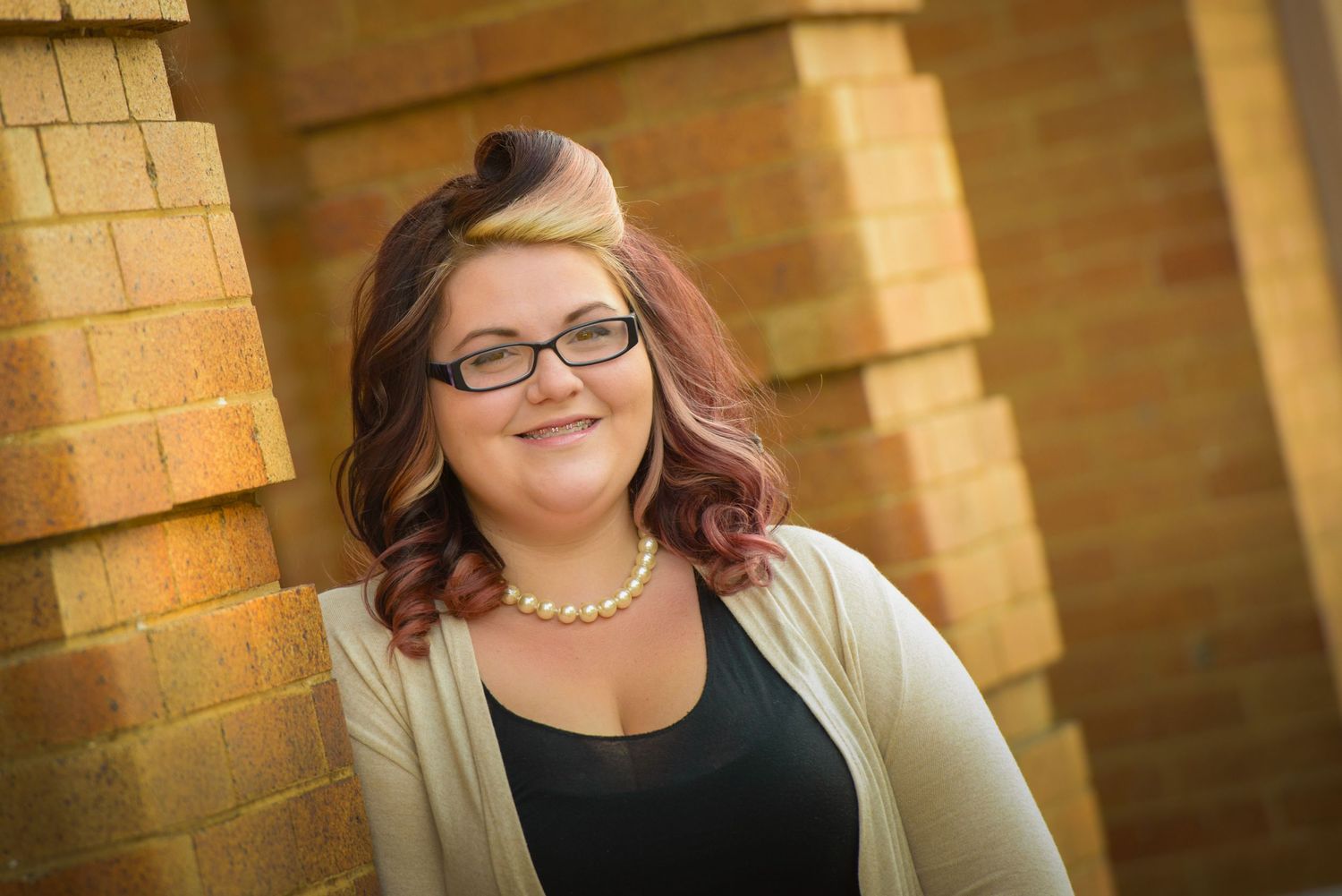 Professional portrait against a brick wall with soft lighting and warm tones, wearing glasses and a cardigan.