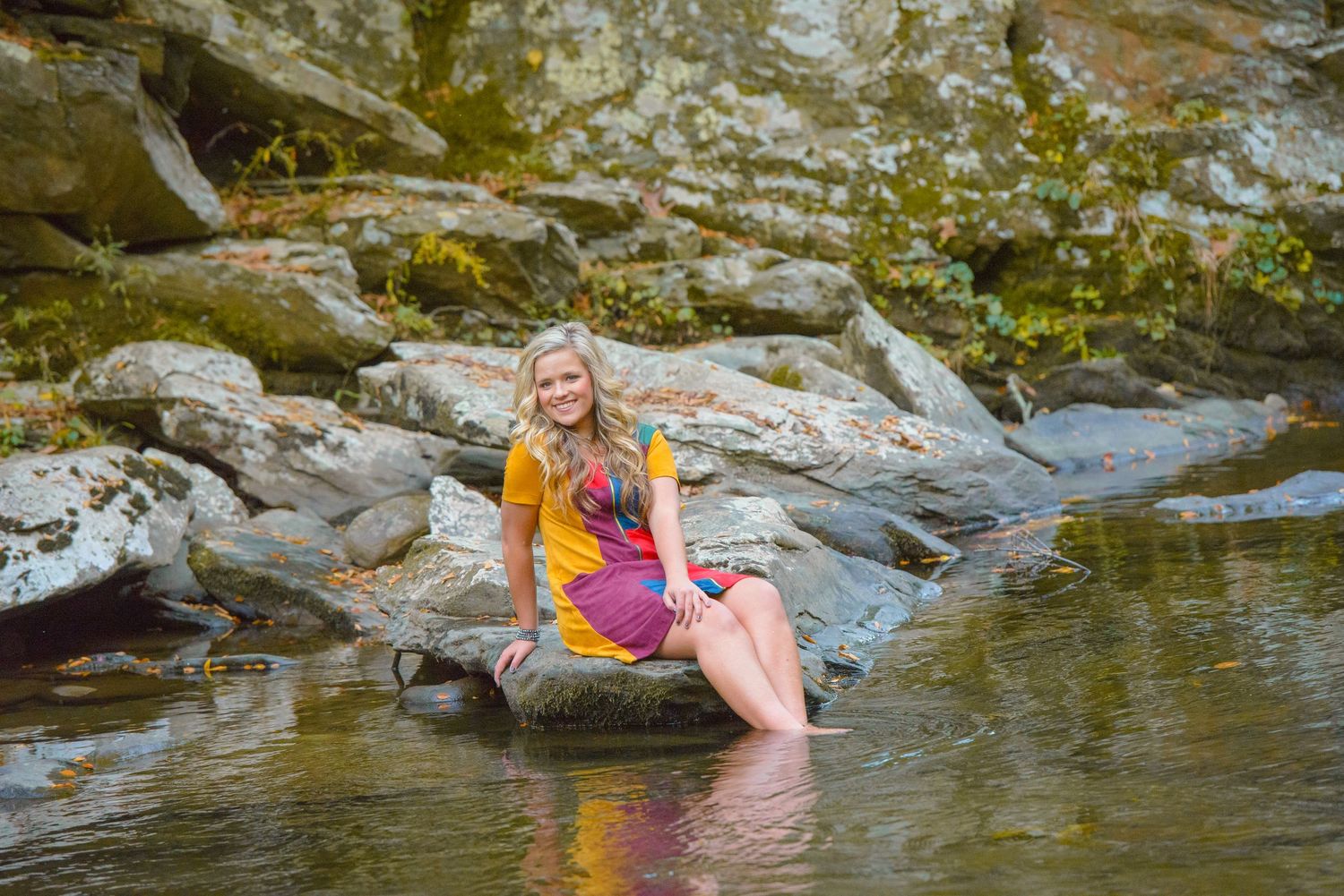 A colorful figure sits on a rock in a stream wearing a yellow and pink outfit during a summer day.