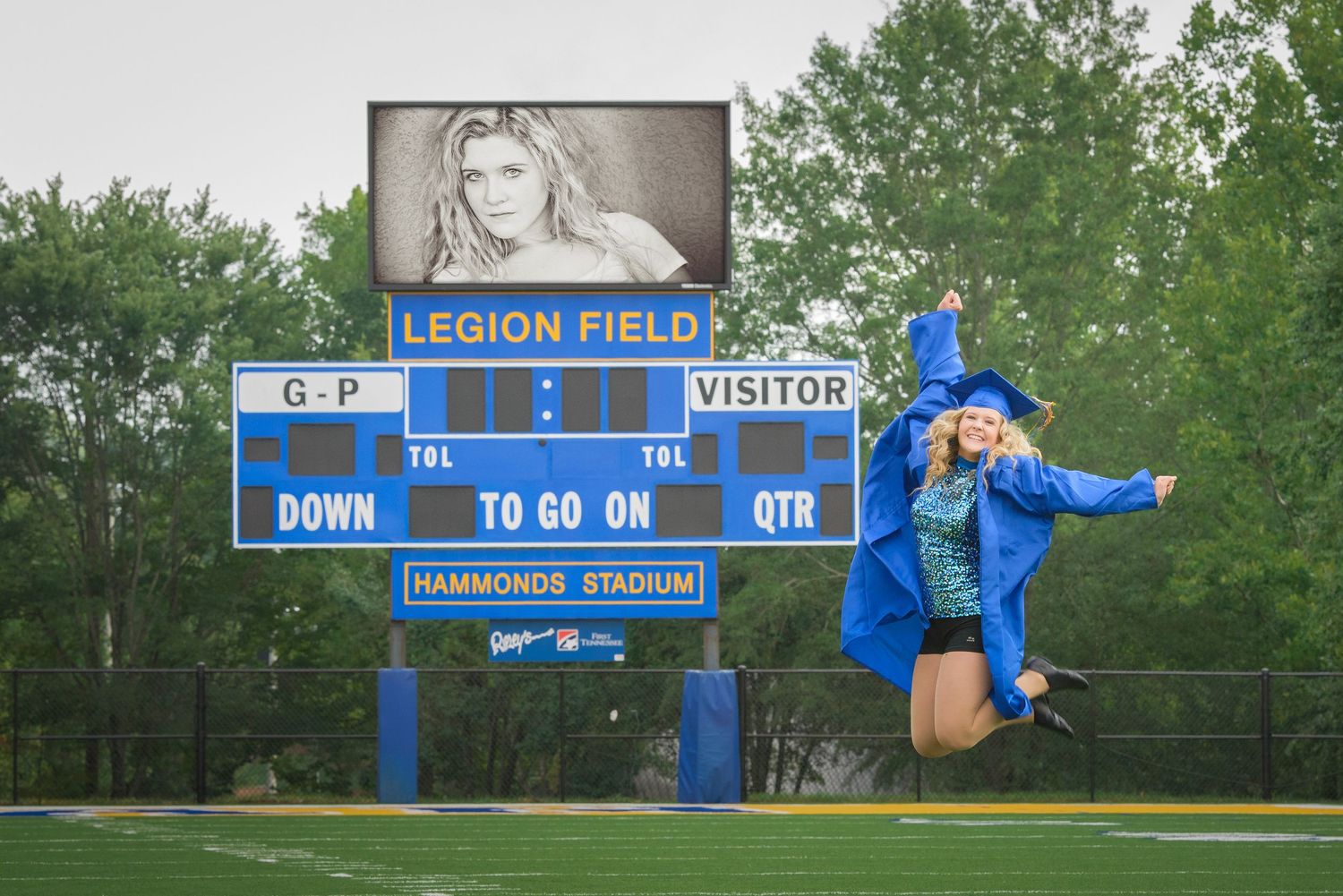 Graduate in blue cap and gown jumps joyfully in front of a Legion Field scoreboard at a sports field during graduation.