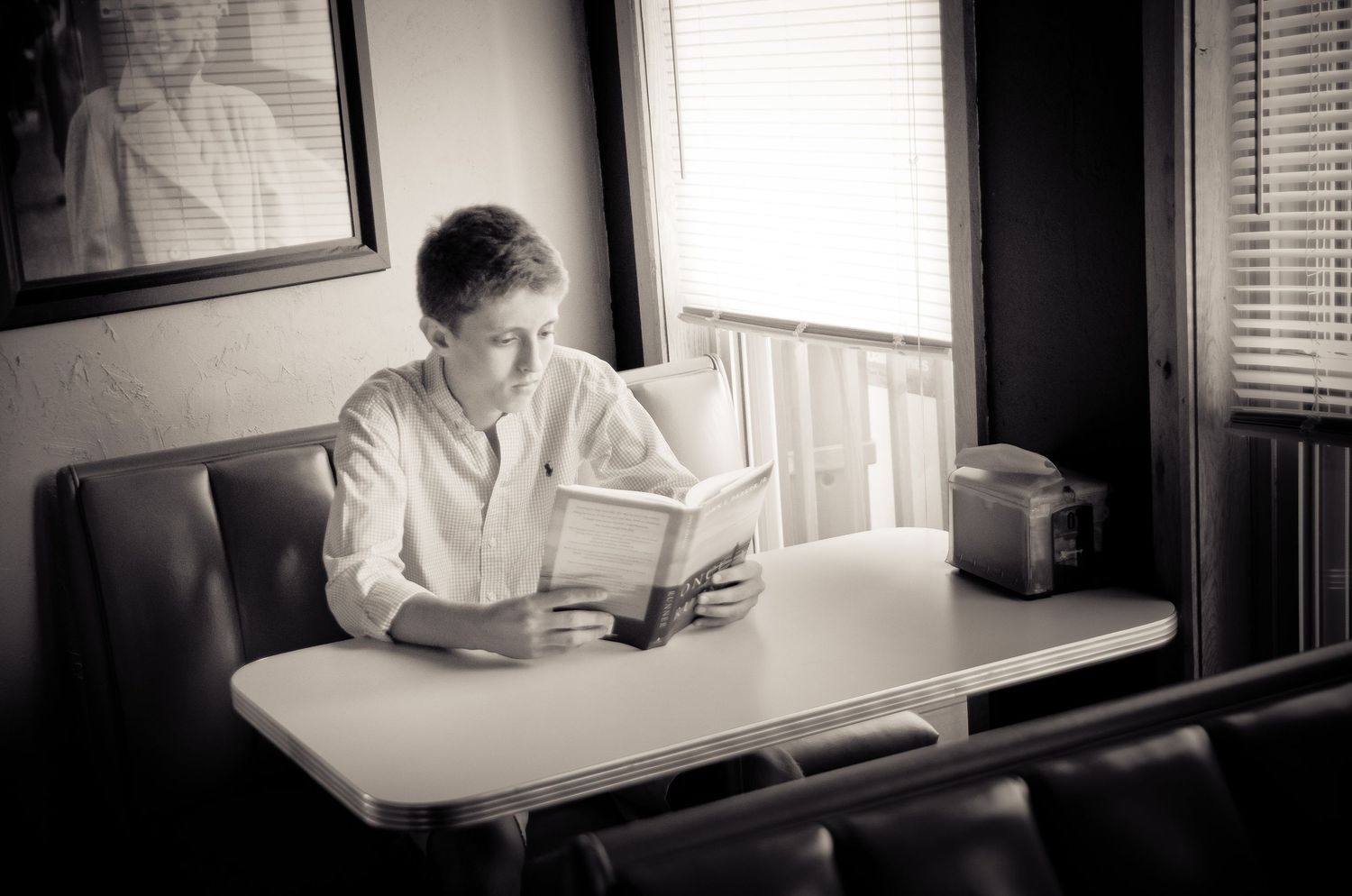 A person in white clothing reads while seated at a table in a dimly lit room with window blinds.