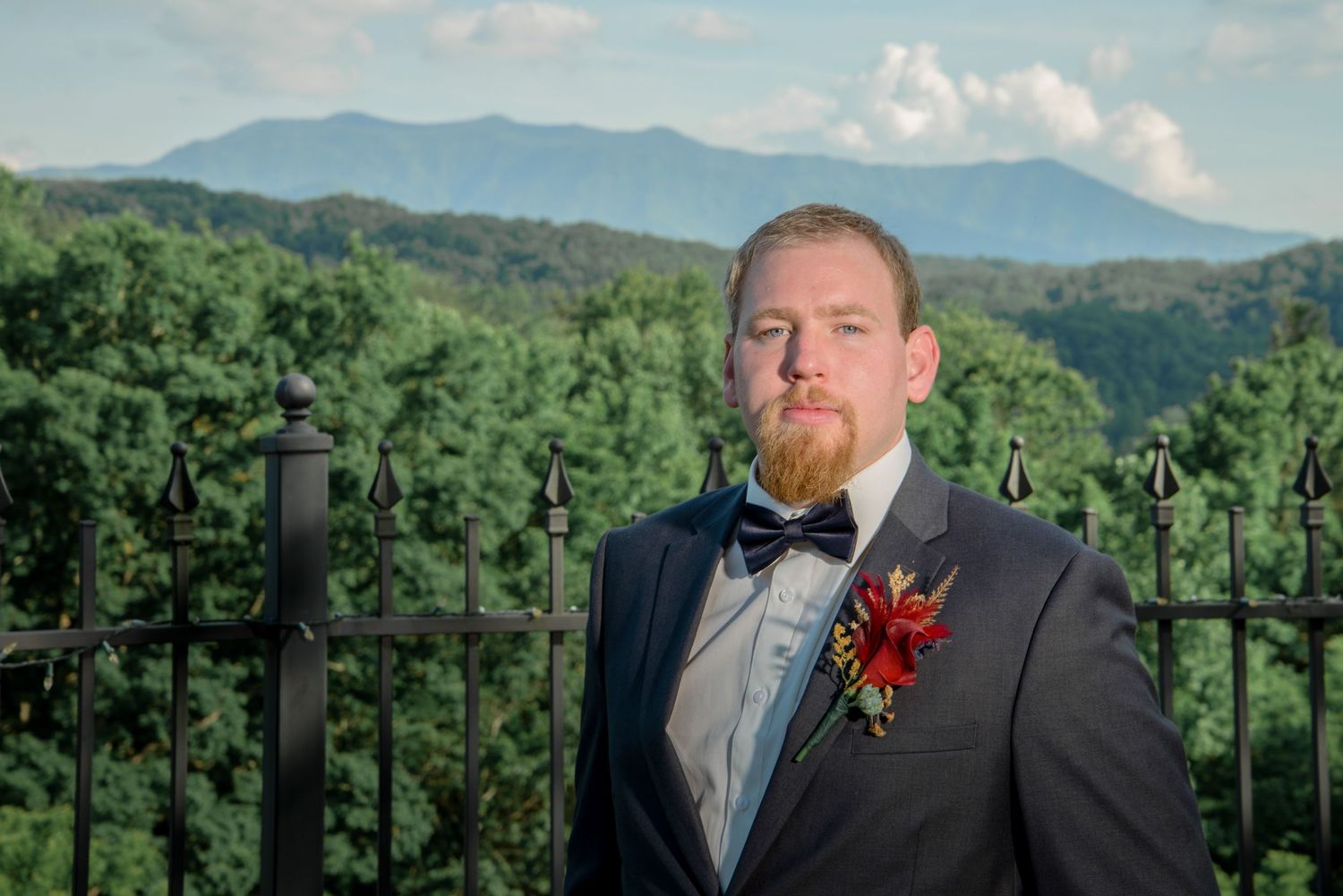 A groom in a black tuxedo with a red boutonniere poses against a scenic mountain backdrop with lush greenery in Pigeon Forge.
