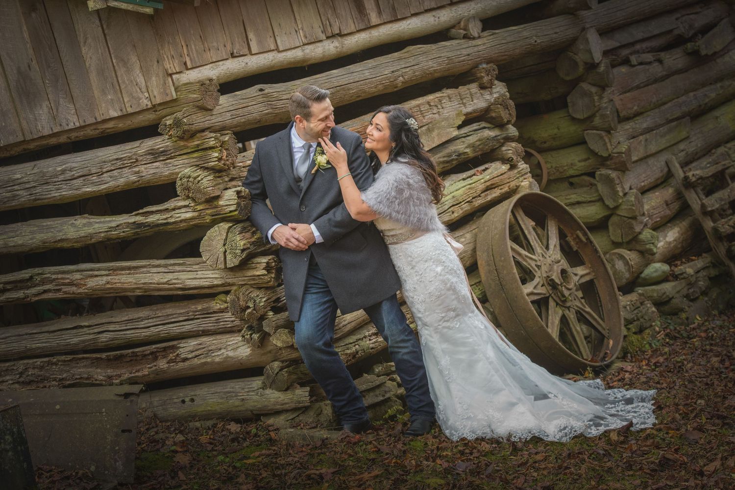 A couple in formal wedding attire pose together against a rustic log wall backdrop with vintage wagon wheel in Pigeon Forge.