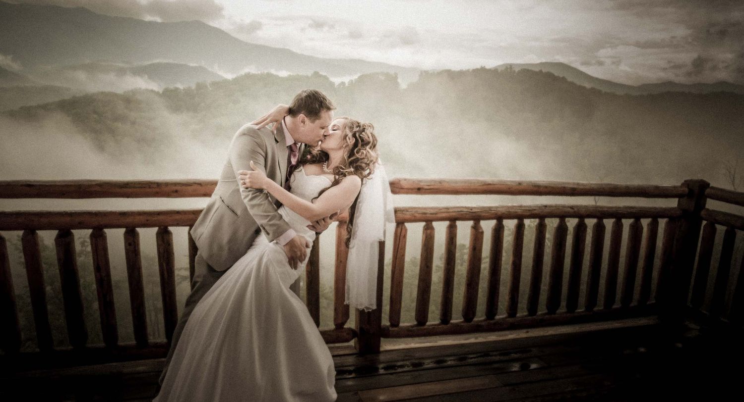 A romantic couple in wedding attire embrace on a misty mountain overlook with wooden railing at sunset.