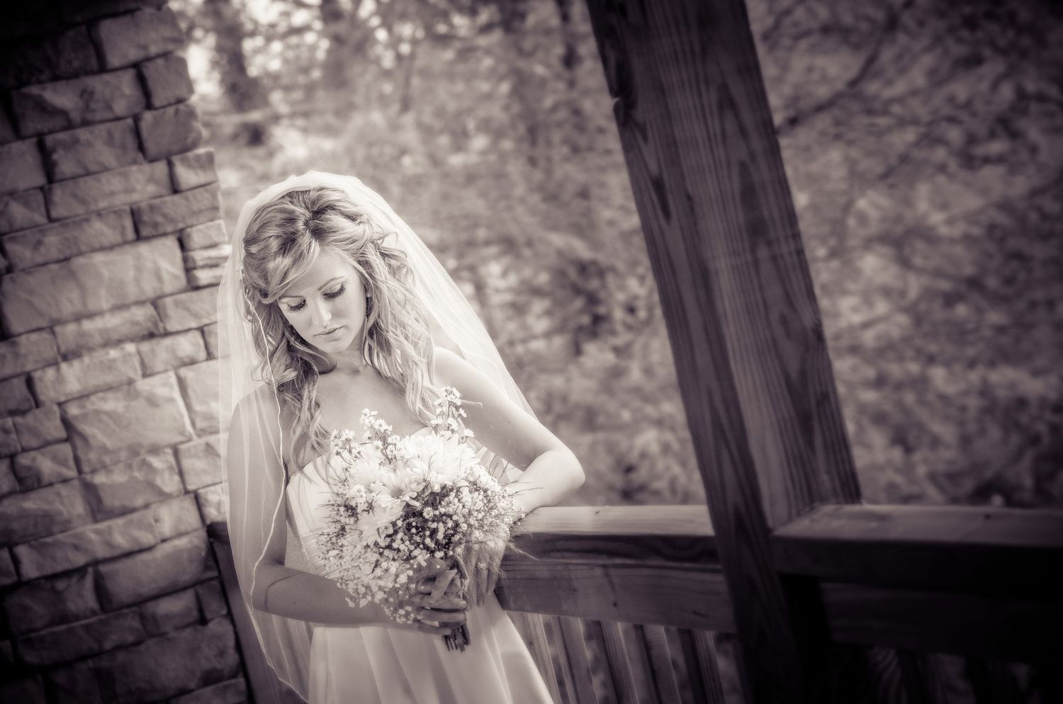 A bride in a traditional white gown and veil holds a bouquet while standing near a rustic brick and wooden structure by Gatlinburg Weddings.