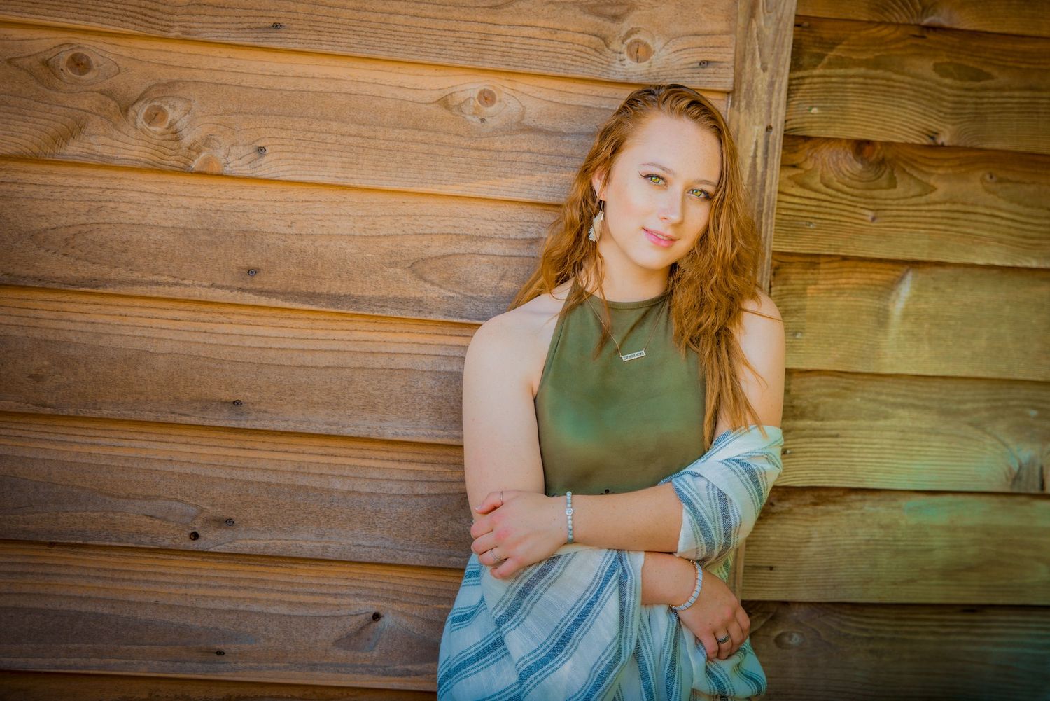 Person in olive green tank top and blue skirt poses against rustic wooden wall in warm sunlight.