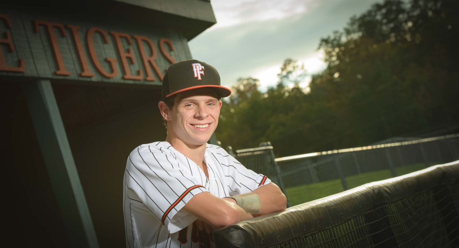 High School Baseball player in uniform leaning against dugout wall at sunset.
