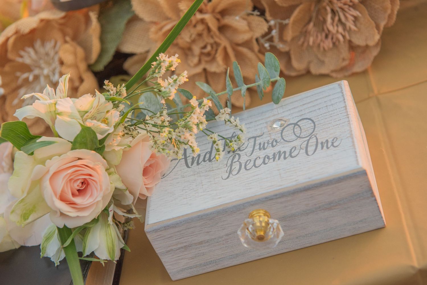 Rustic wooden box with 'And Two Became One' text next to pink roses and white floral arrangements.