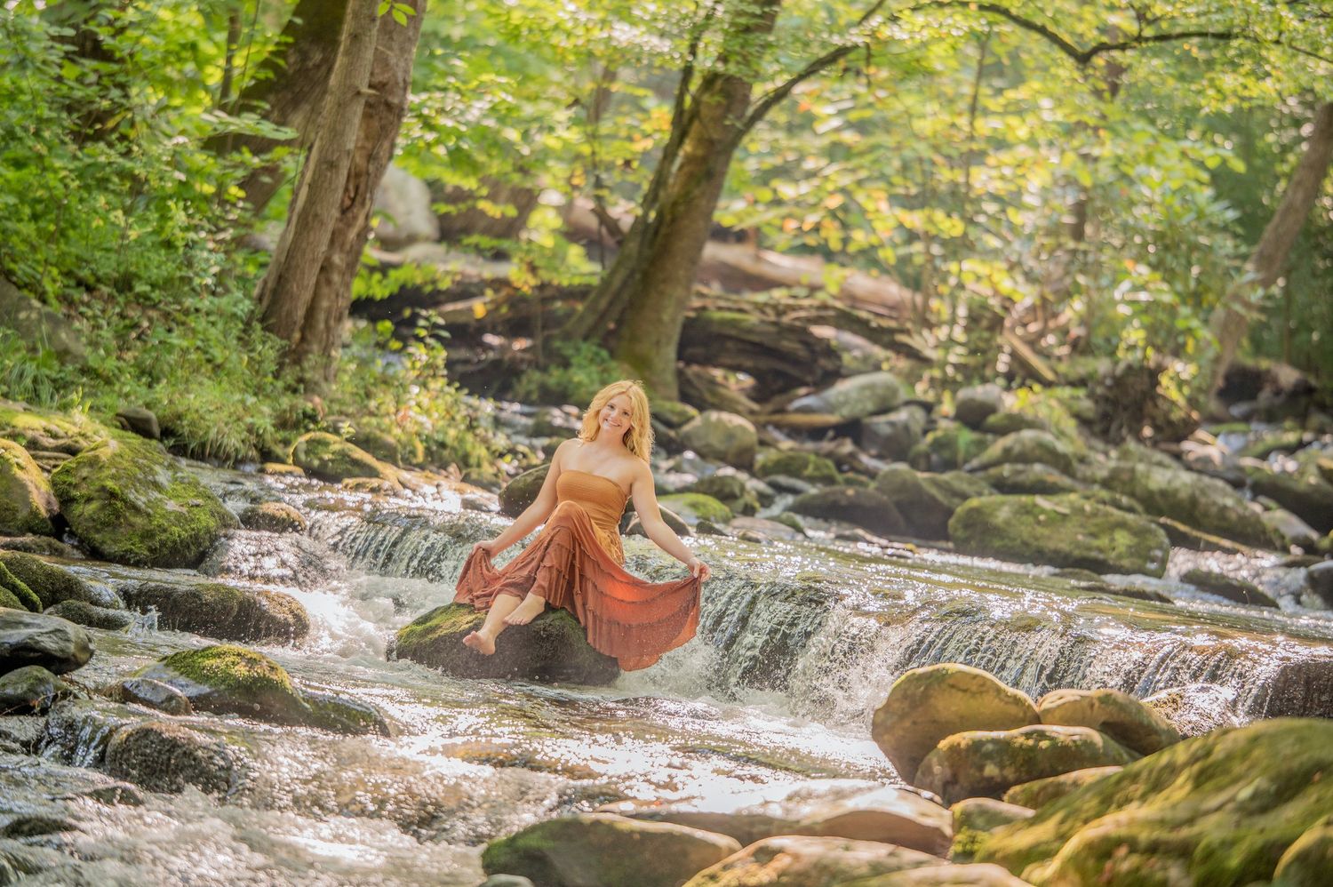 Gatlinburg Senior pictures best photograph. Girl in orange dress sitting on rocks by a flowing forest stream surrounded by lush green trees.