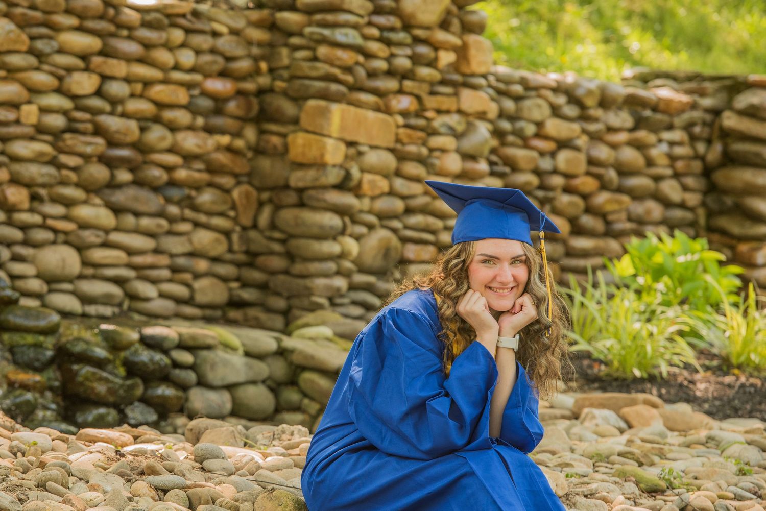 Graduate in bright blue cap and gown sits thoughtfully by rustic stone wall with garden backdrop.
