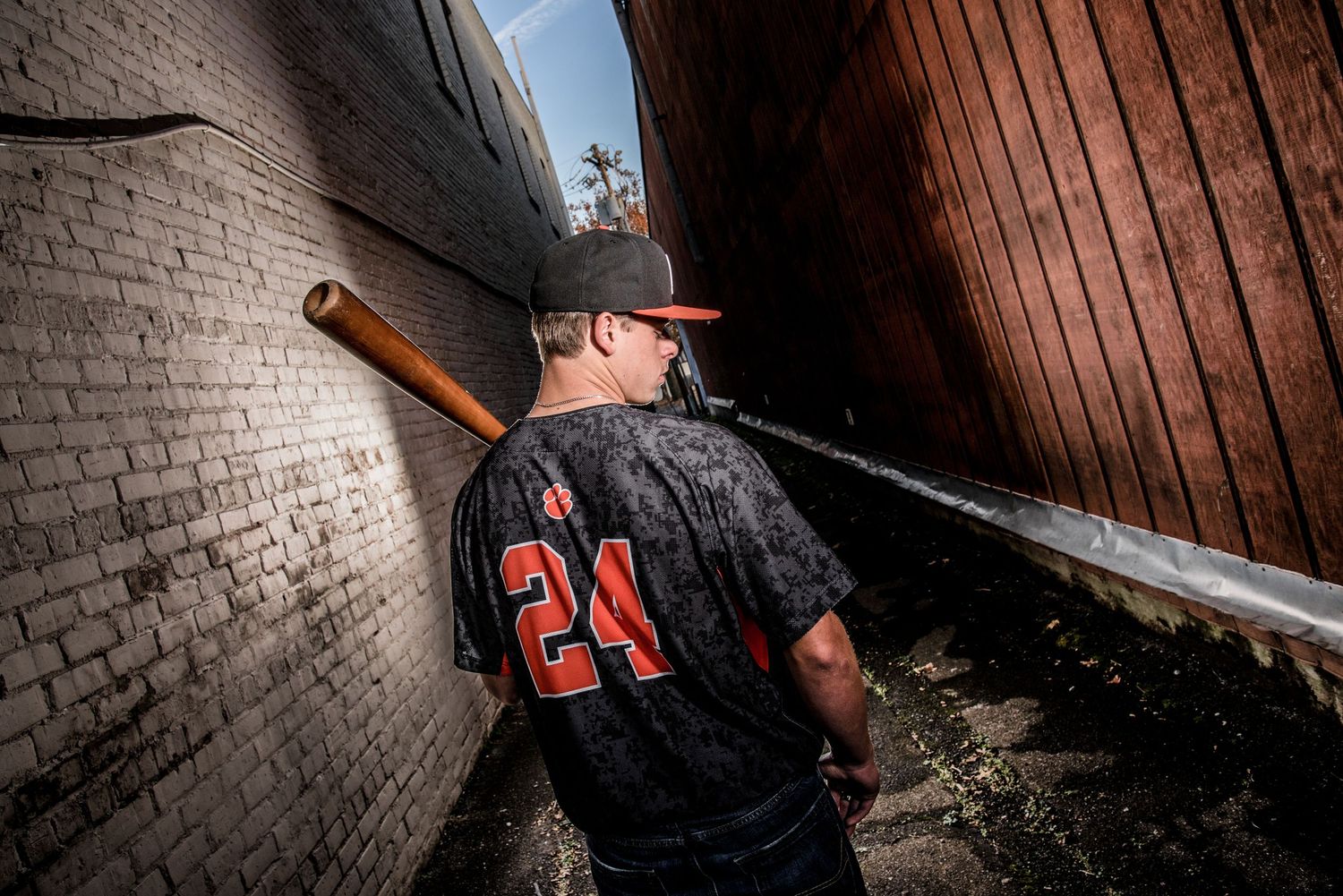 Baseball player in black jersey number 24 stands in an alley between brick walls with bat over shoulder.
