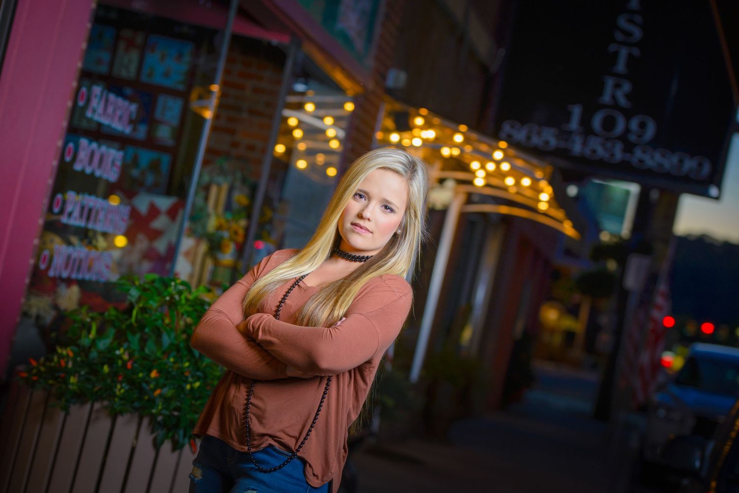 A person stands with crossed arms in front of a storefront at night with twinkle lights in the background.