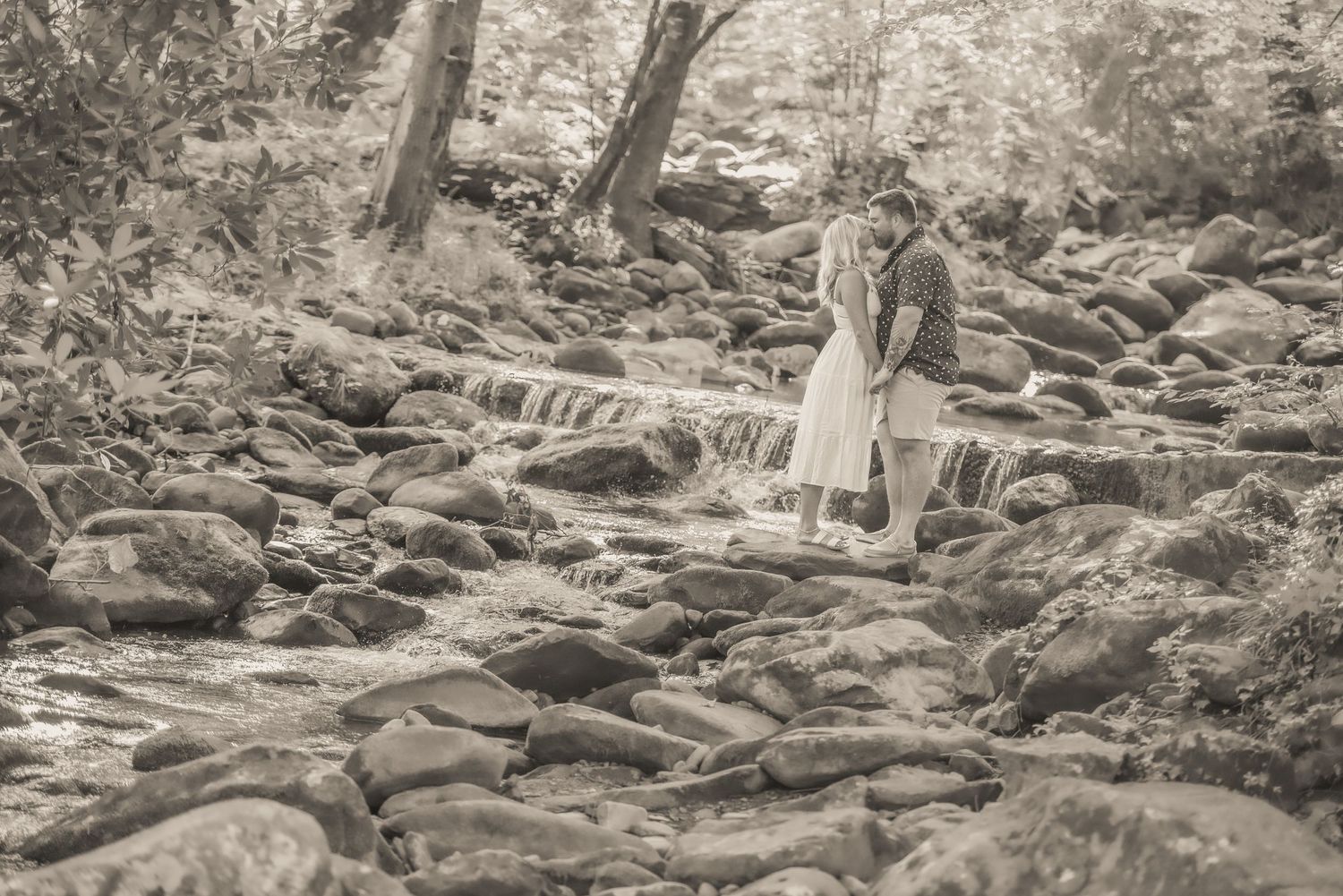 A romantic black and white photo of a couple standing on rocks near a flowing stream in a wooded forest setting by gatlinburg photographers.