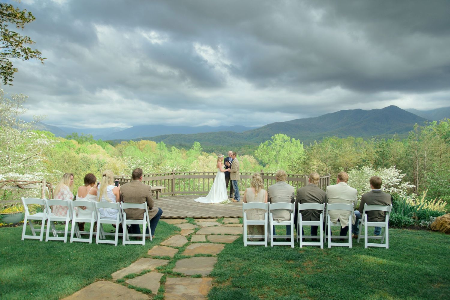 A small outdoor wedding ceremony takes place on a mountain overlook with dramatic storm clouds and green scenery.