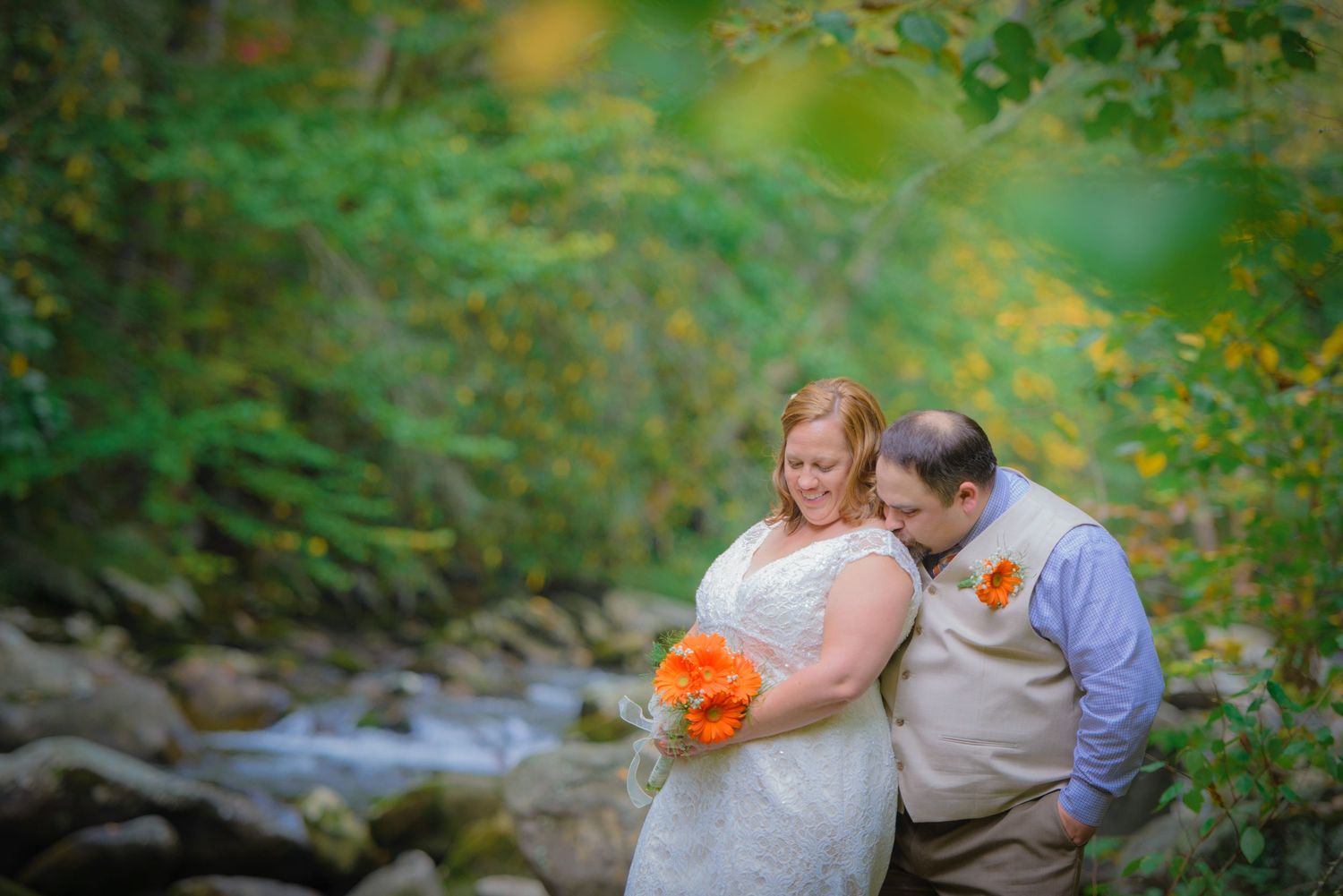 A couple embraces in a dreamy forest setting with orange flowers and a flowing stream in the background in Pigeon Forge TN.