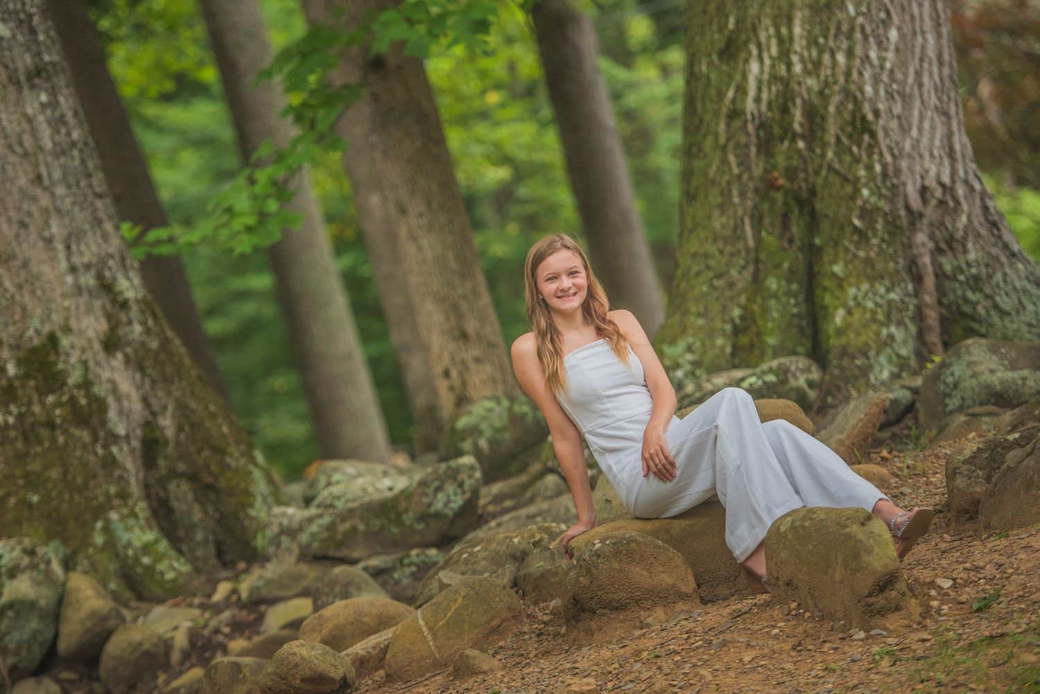 Figure in a white dress reclines on a fallen log in a lush forest setting during golden hour lighting.