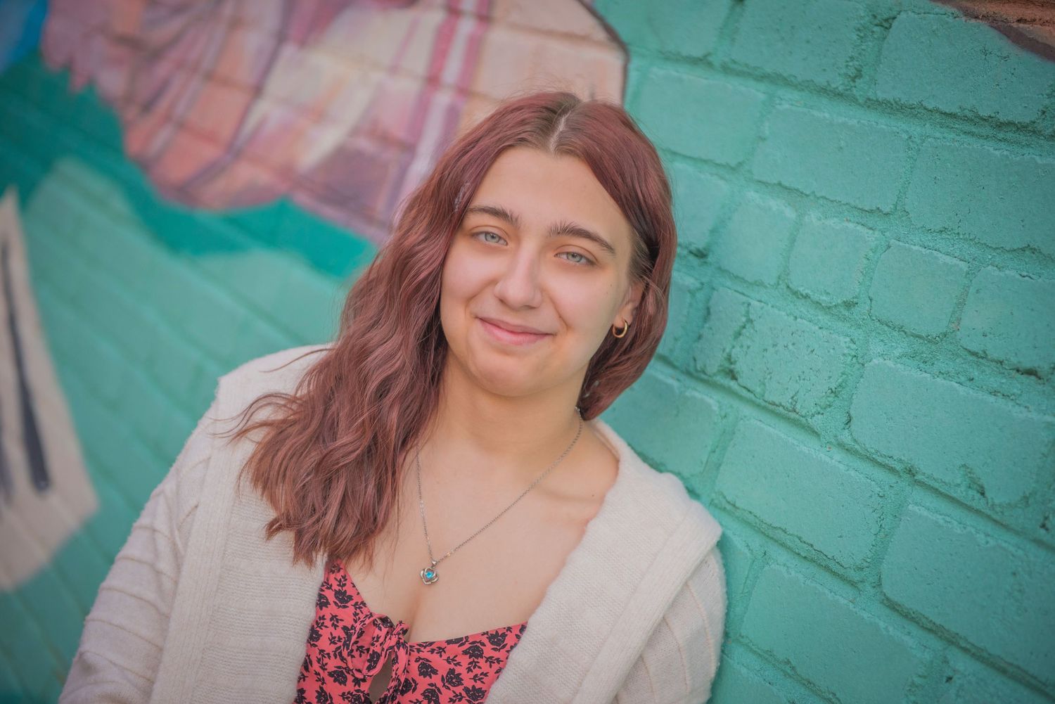 A smiling person wearing a cardigan stands against a bright turquoise brick wall in soft, warm lighting.