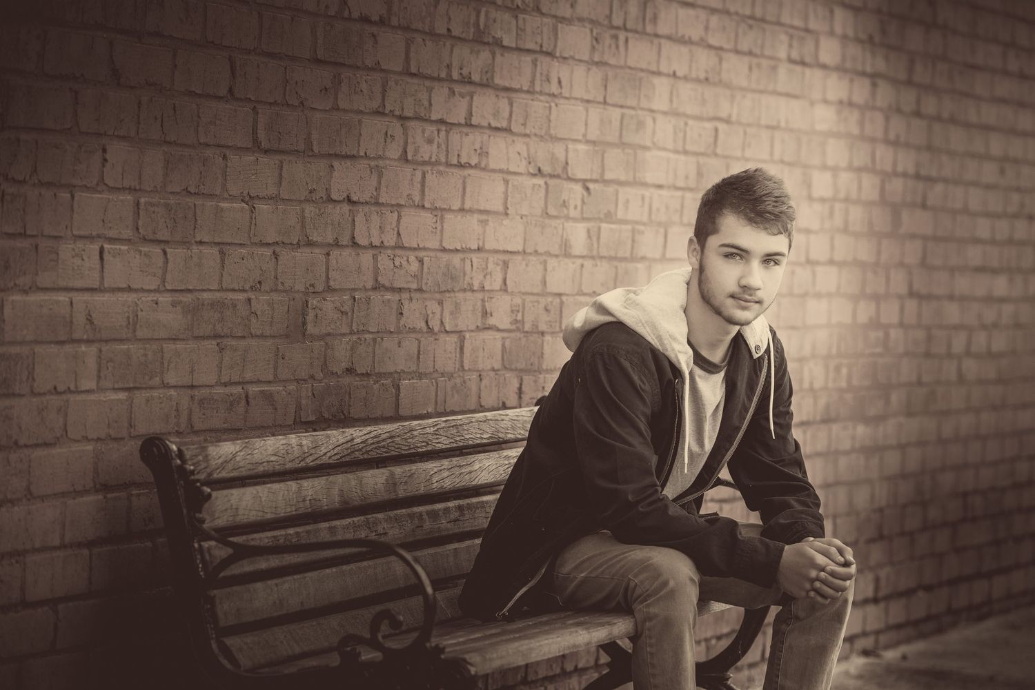 Black and white portrait of a person sitting on a wooden bench against a brick wall in vintage style lighting.