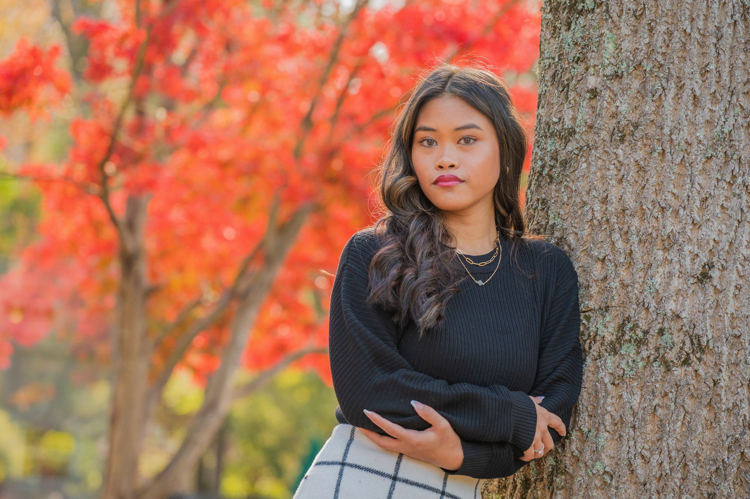 Young woman in a black sweater and plaid skirt stands near tree with vibrant red autumn foliage in background.