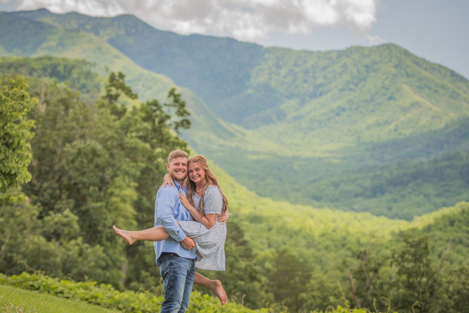 A couple embraces while standing on a hilltop overlooking lush green mountains on a summer day by Gatlinburg TN Photographers.