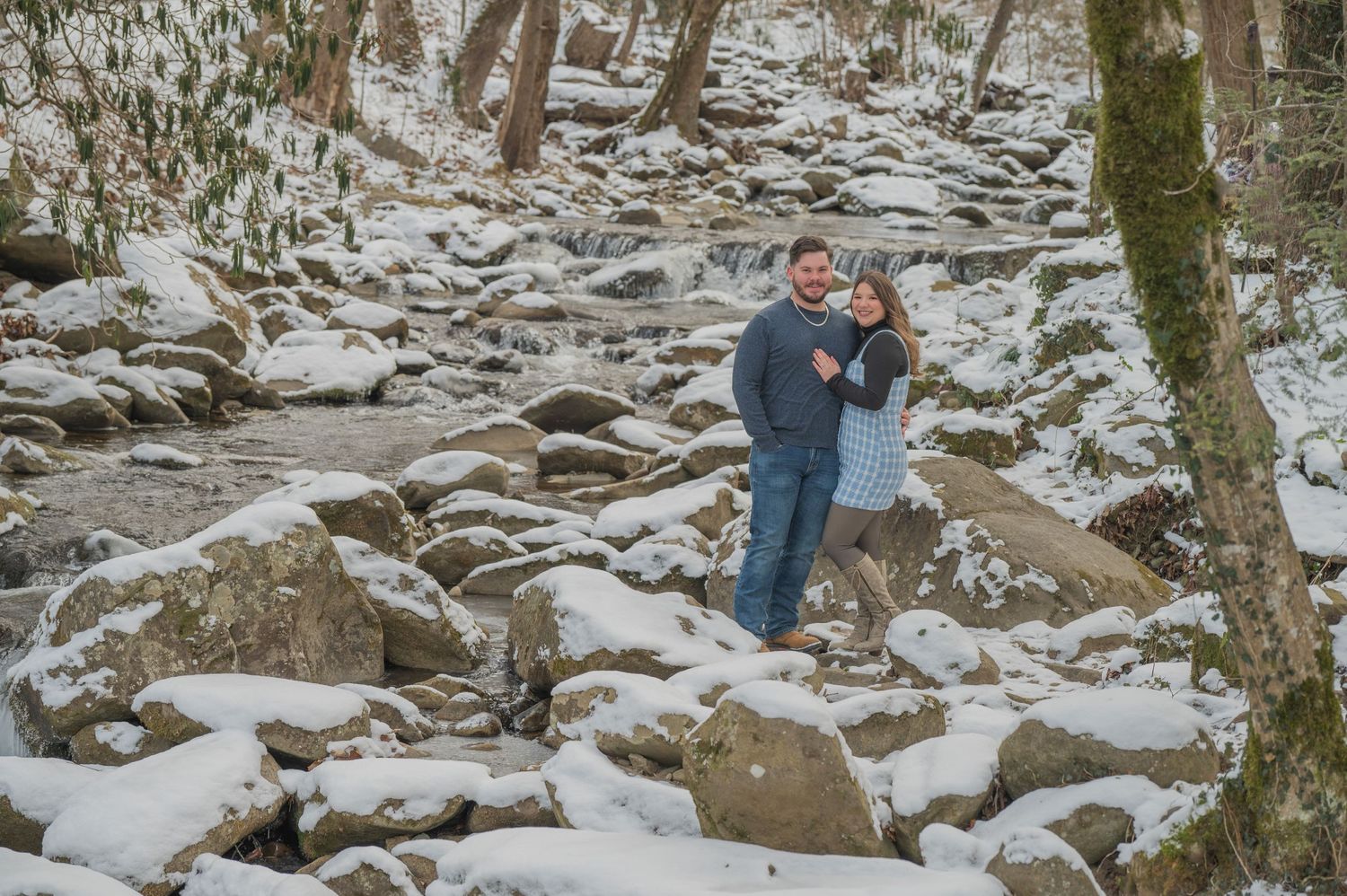 Two people embrace while standing among snow-covered rocks in a winter forest setting by Pigeon Forge Photographers tn.