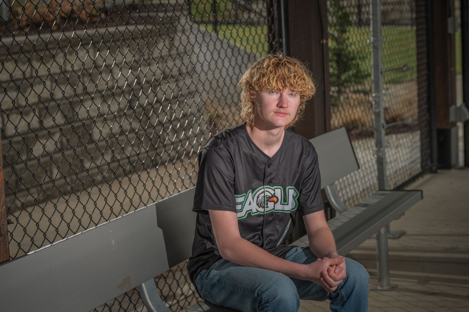 A student wearing a dark UCLA t-shirt sits on a bench beside a chain link fence during golden hour lighting by Pigeon Forge Photographers tn