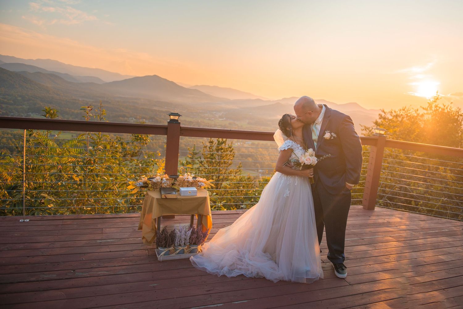 A romantic wedding moment captured on a deck overlooking mountains at sunset with yellow wildflowers in the foreground.