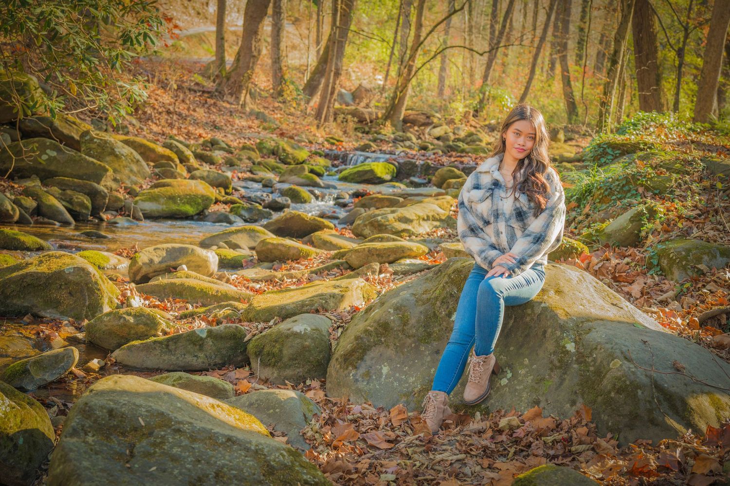 Someone sits on moss-covered rocks by a stream in an autumn forest with golden leaves scattered on the ground.