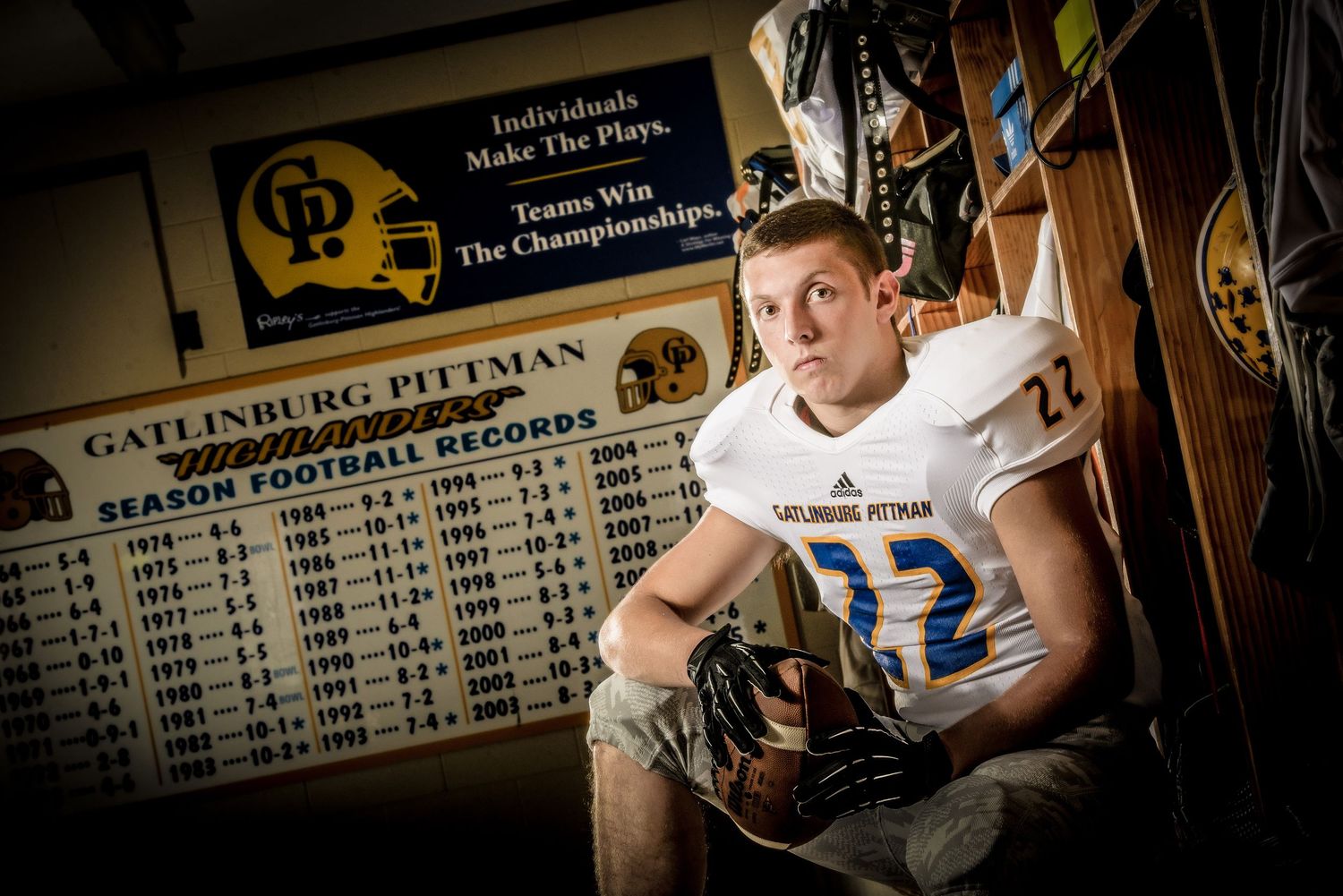 A football player in white uniform sits in a locker room beneath a statistics board and sports memorabilia.