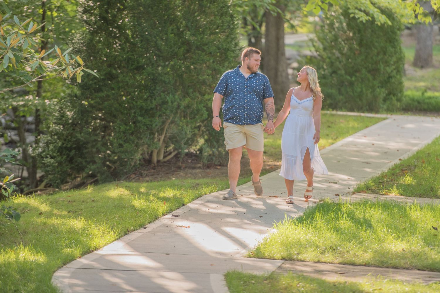 A engagement couple strolls together on a sunny garden path surrounded by lush green landscaping and trees by Sevierville TN photographer.