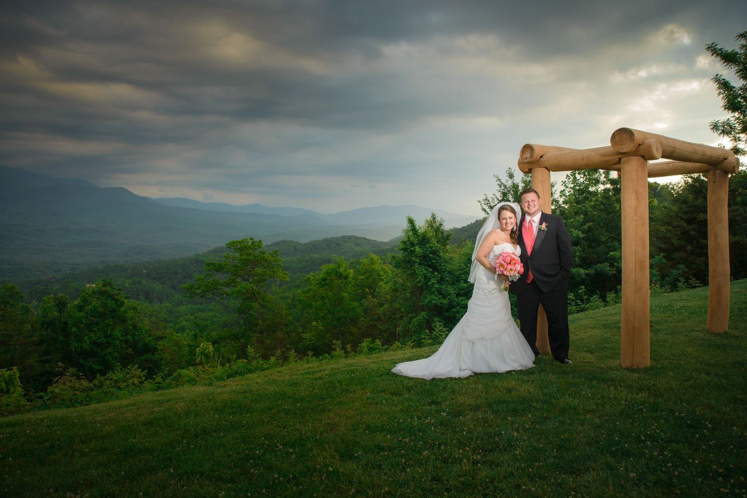 A couple embraces under a wooden arch atop a mountain overlook with dramatic cloudy skies and rolling hills beyond in Gatlinburg TN.