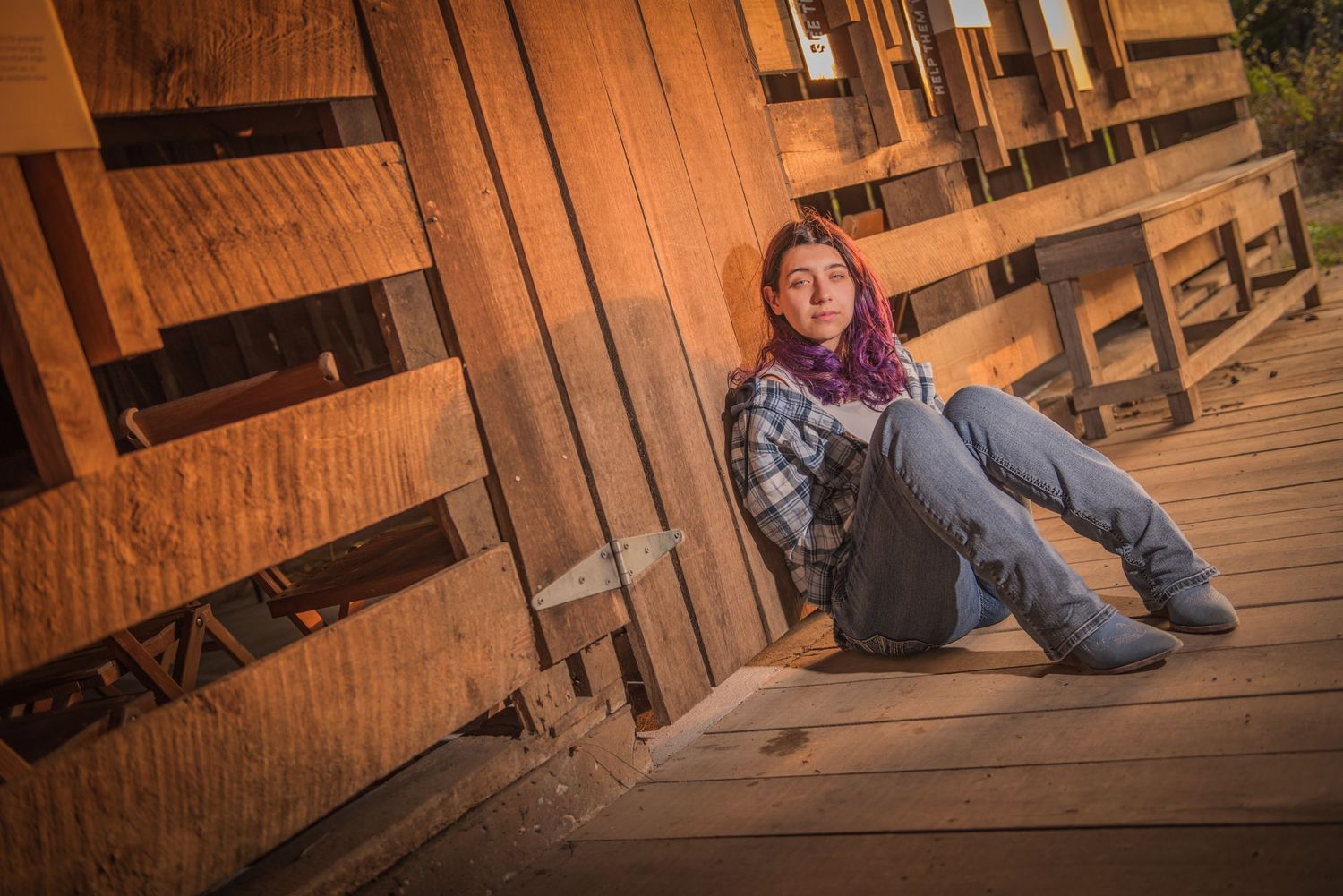 Person with colorful hair sitting casually against wooden wall on rustic deck in warm evening light.