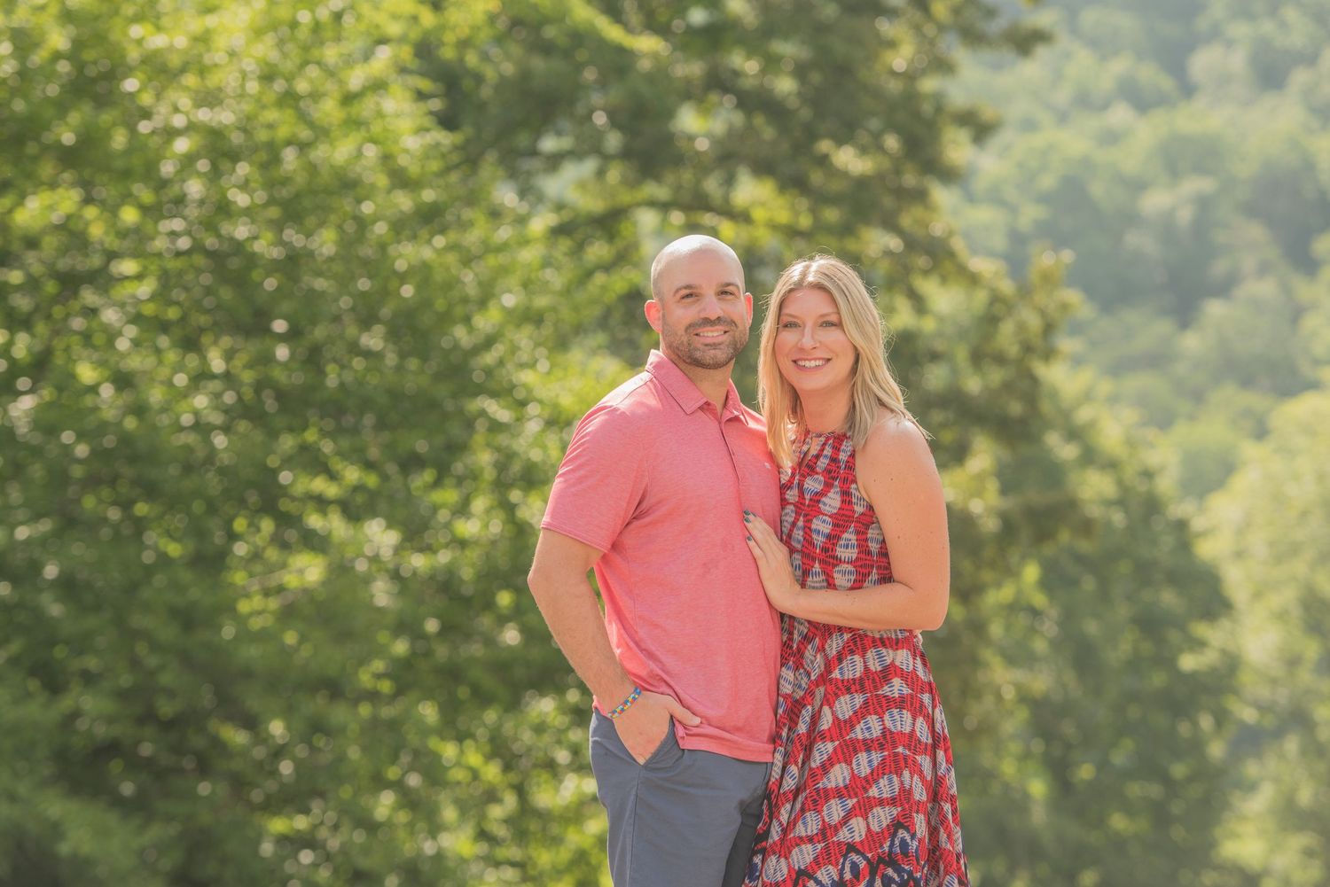 An engagement couple stands together outdoors, with a person wearing coral pink and another in a patterned red dress against lush greenery.