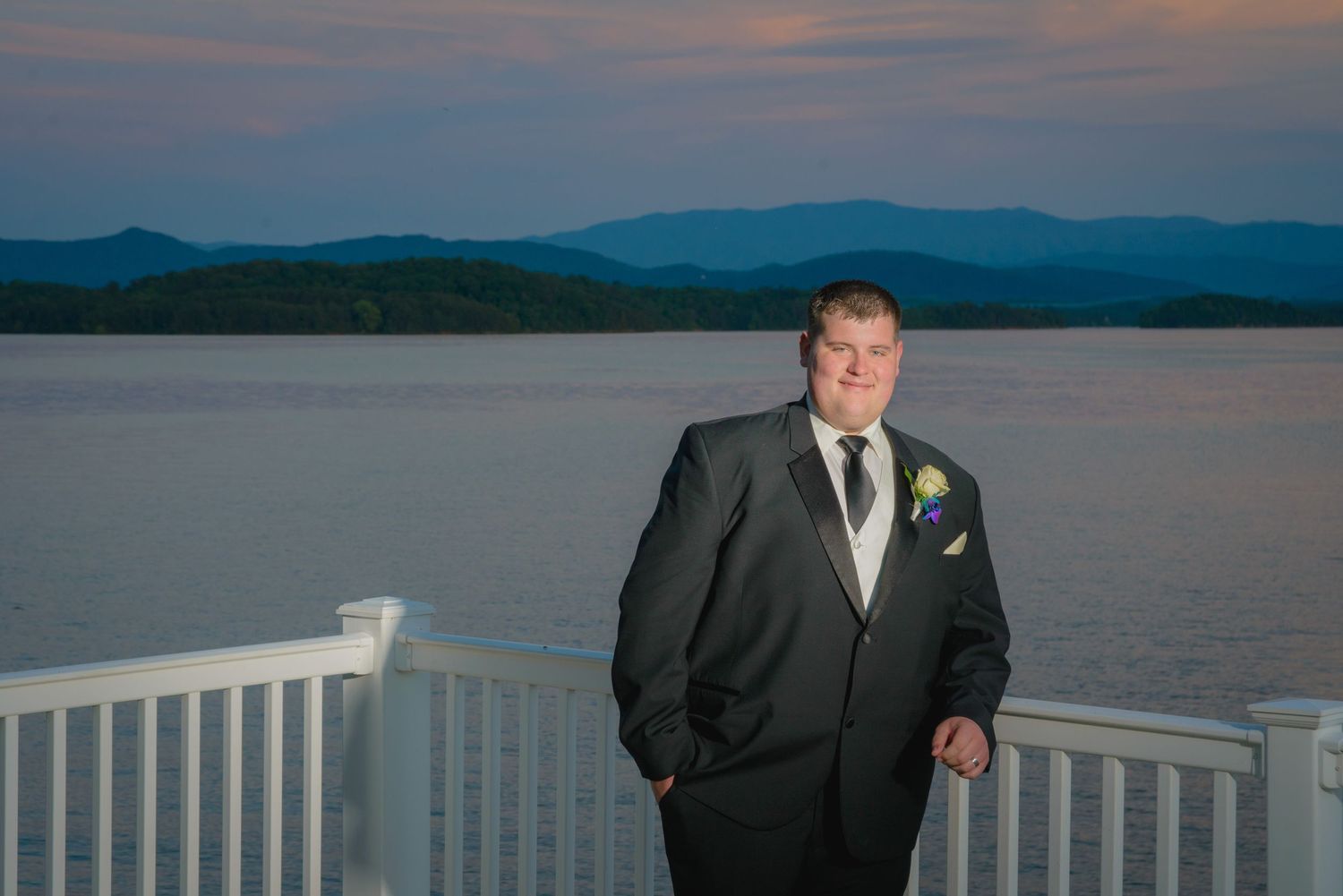 A groom in a black suit stands on a deck overlooking a scenic lake and mountain landscape at dusk by Gatlinburg Wedding Photographers.
