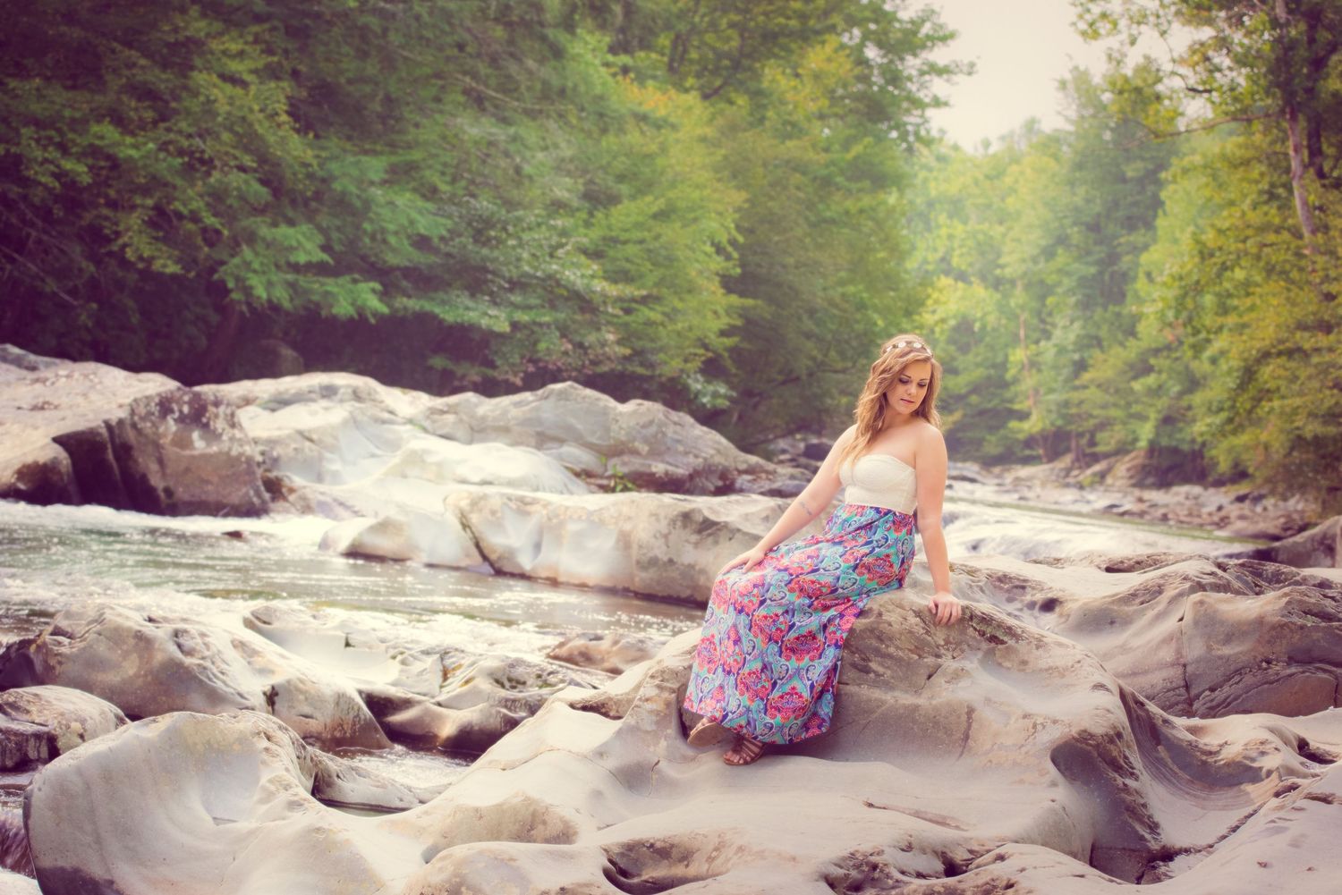A person in a floral maxi dress sits gracefully on rocky river terrain surrounded by lush forest scenery.