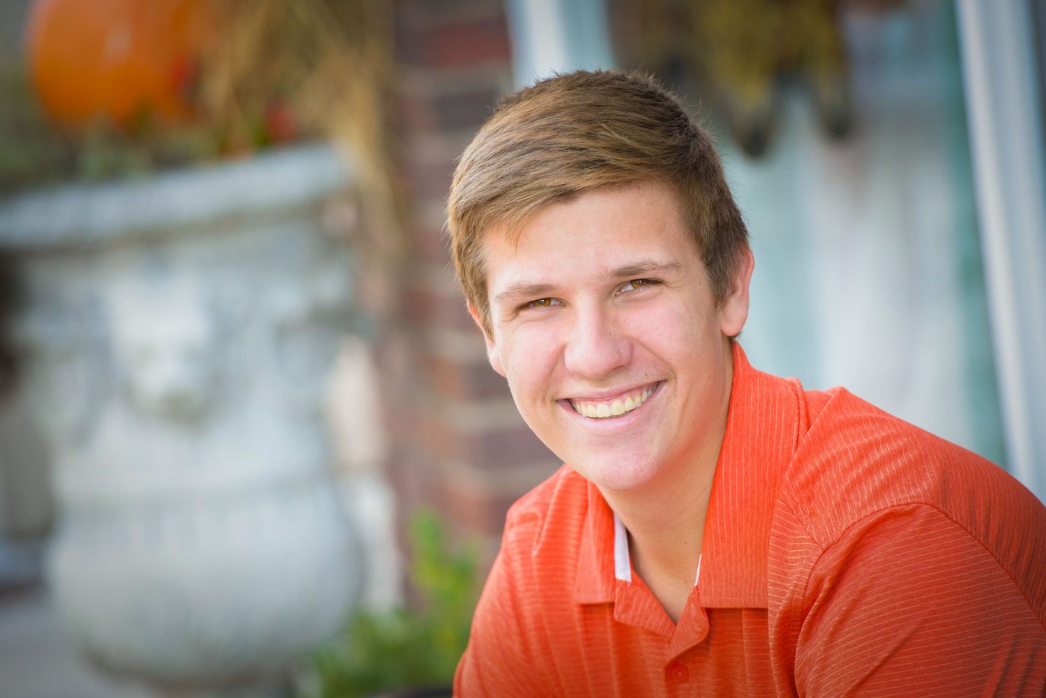 A person in a bright orange polo shirt smiles warmly while sitting outdoors with autumn decorations in the background.