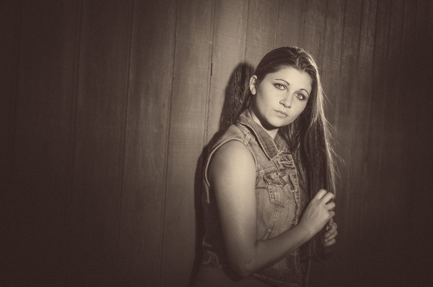 A sepia toned portrait of a woman in a floral dress against a wooden wall background.