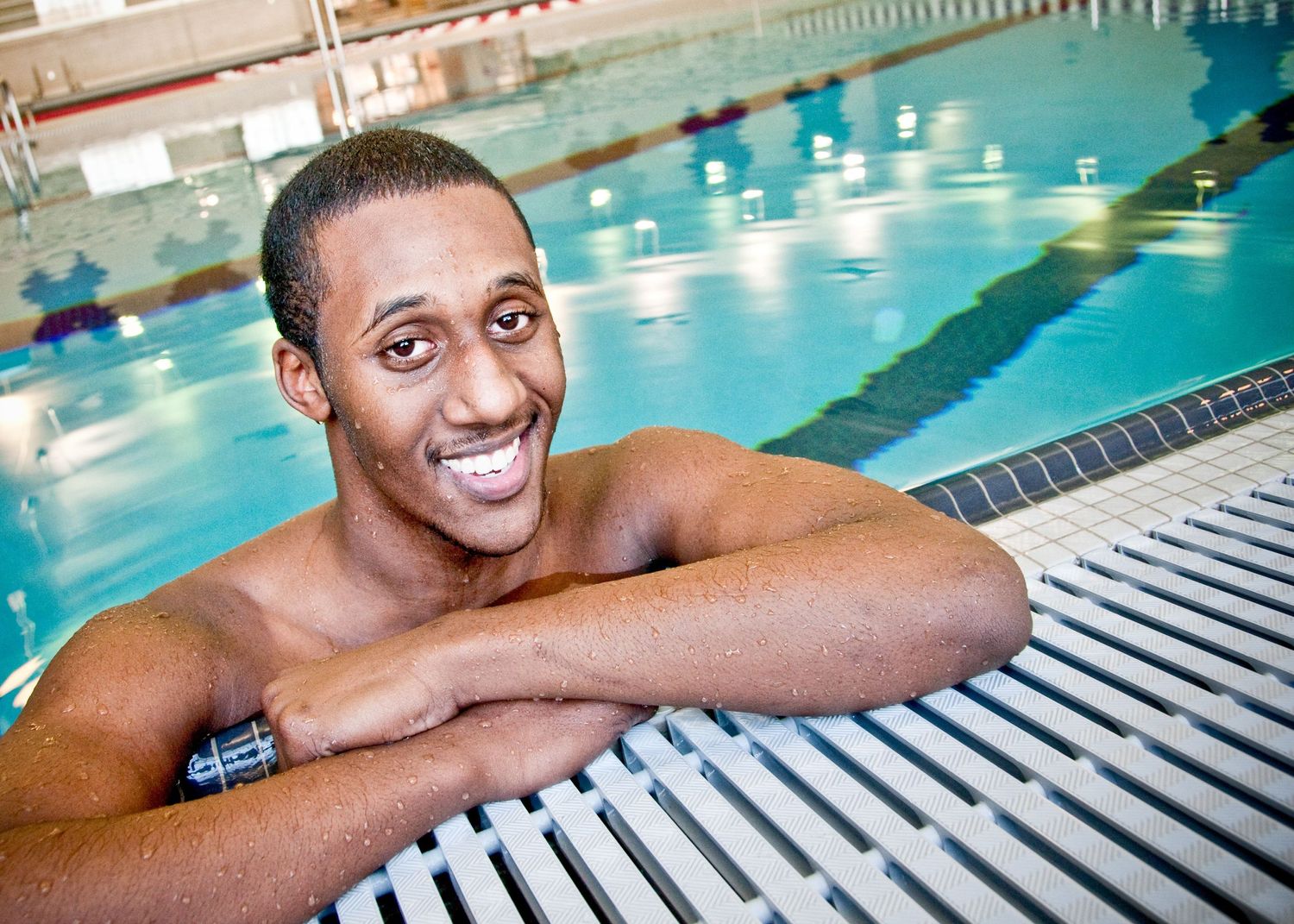 Swimmer rests arms on pool deck edge with bright smile while in indoor swimming pool with lane lines.