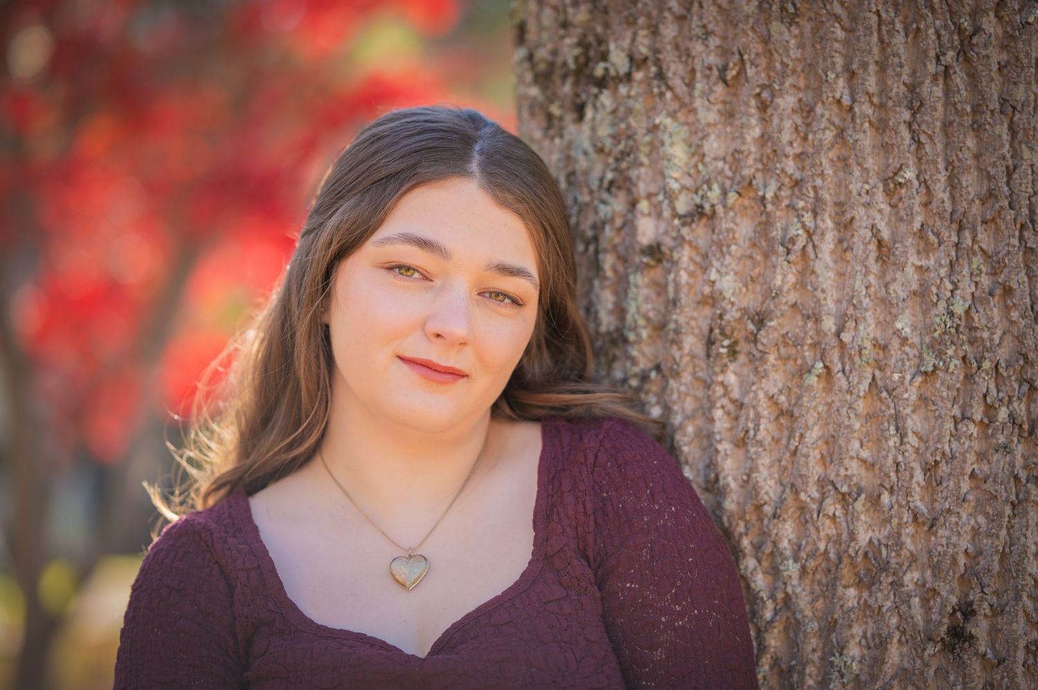 Portrait of someone in a burgundy sweater leaning against a tree trunk with red autumn foliage in the background.
