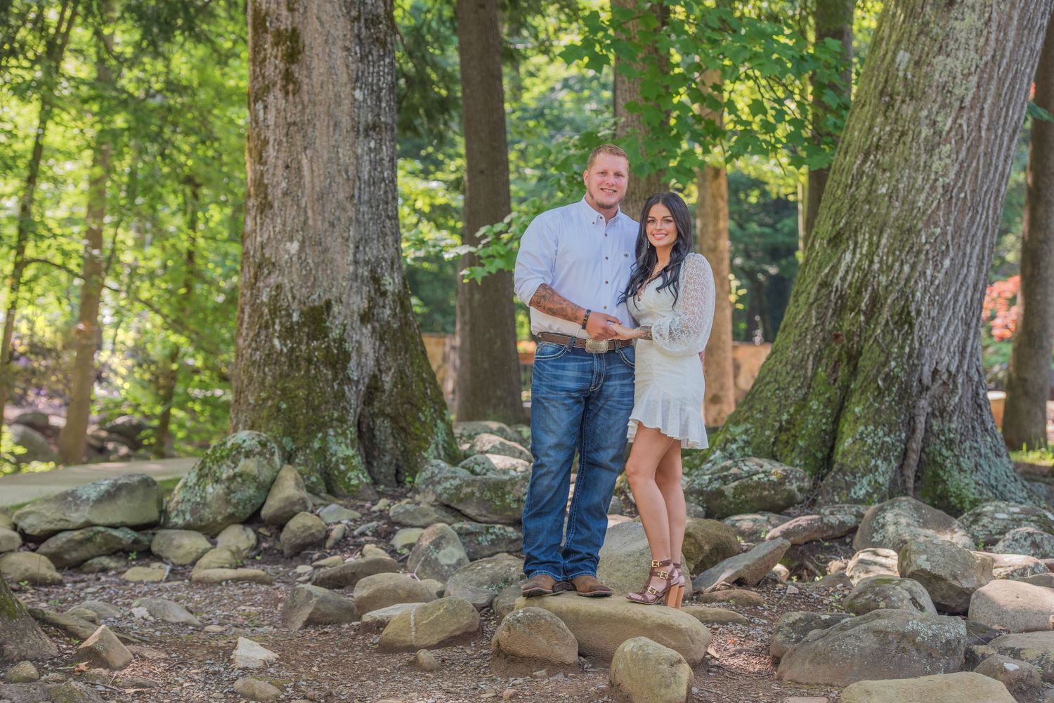An engagement couple stands together among large tree trunks and rocks in a scenic wooded setting on a sunny day in Sevierville TN.