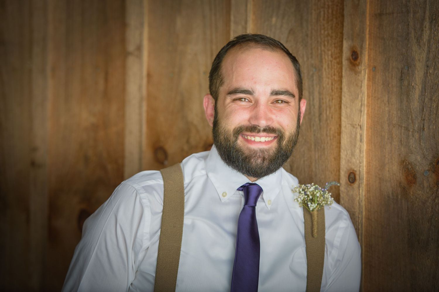 A person with a full beard wearing a white dress shirt, purple tie and tan suspenders stands against a wooden wall in Pigeon Forge TN.