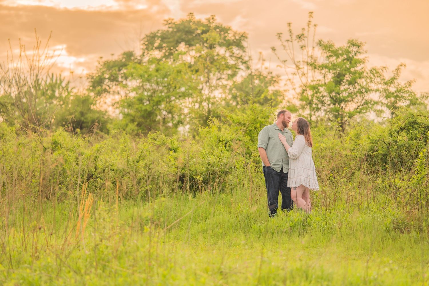 A couple stands close together in a grassy meadow during golden hour with trees in the background by Pigeon Forge Photographer TN,